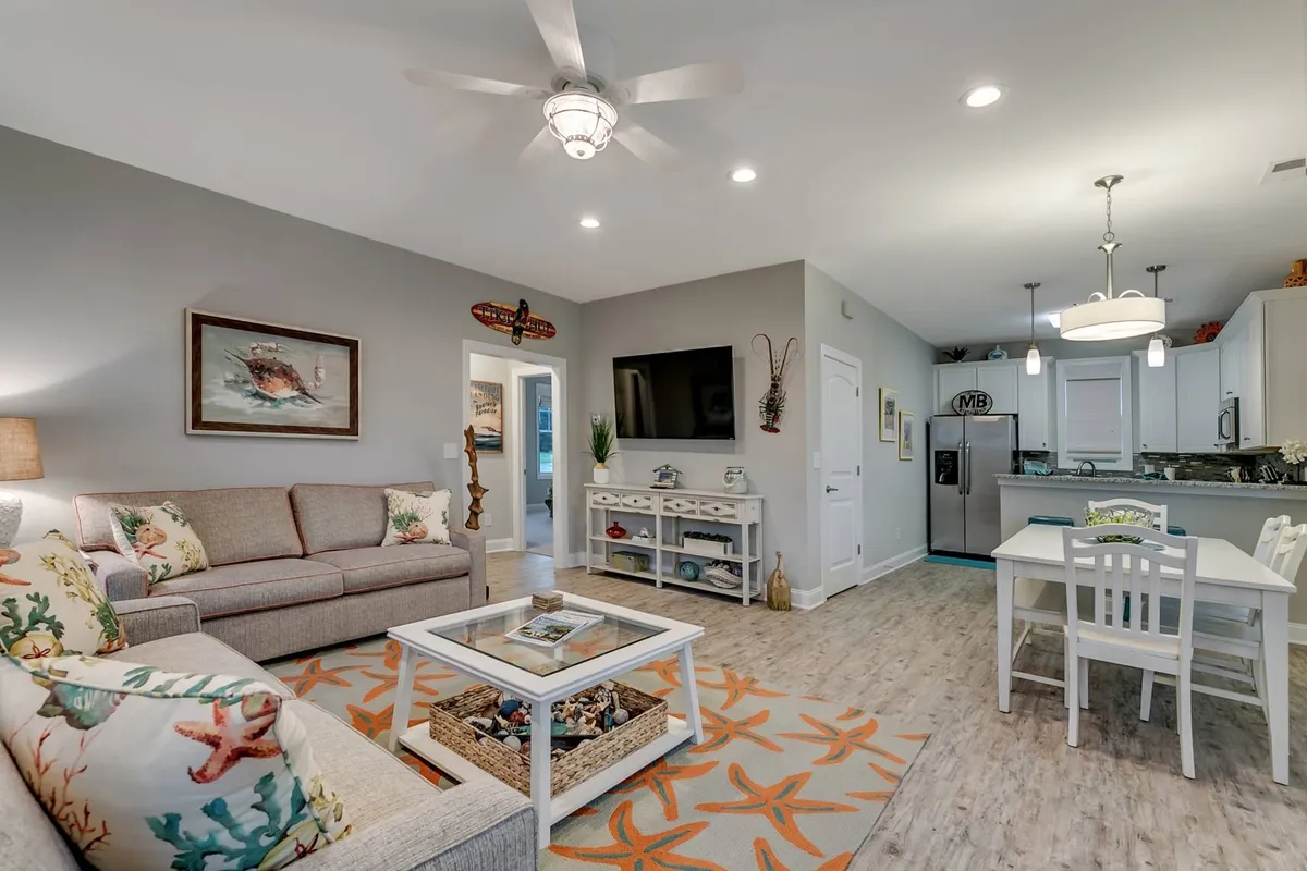A cozy corner of the open living area featuring a coral-patterned sofa, a glass coffee table adorned with shells, a dining table with white chairs, and coastal-inspired wall art. The arrangement showcases how the living space seamlessly blends relaxation with dining and decor.