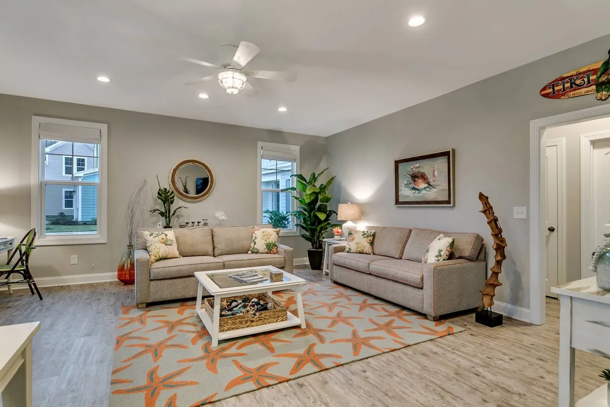 A cozy living room area with a plush sofa adorned with decorative pillows, a coffee table featuring books, and a potted plant. Artwork decorates the wall, and a ceiling fan adds comfort to the inviting atmosphere.