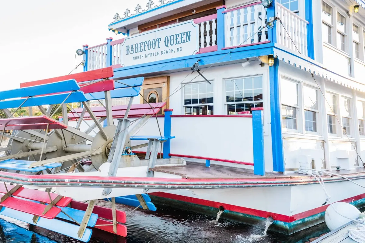 A colorful paddlewheel boat docked near the marina, with vibrant signage and outdoor seating. The scene hints at waterfront dining, boat tours, and a lively, playful resort lifestyle.