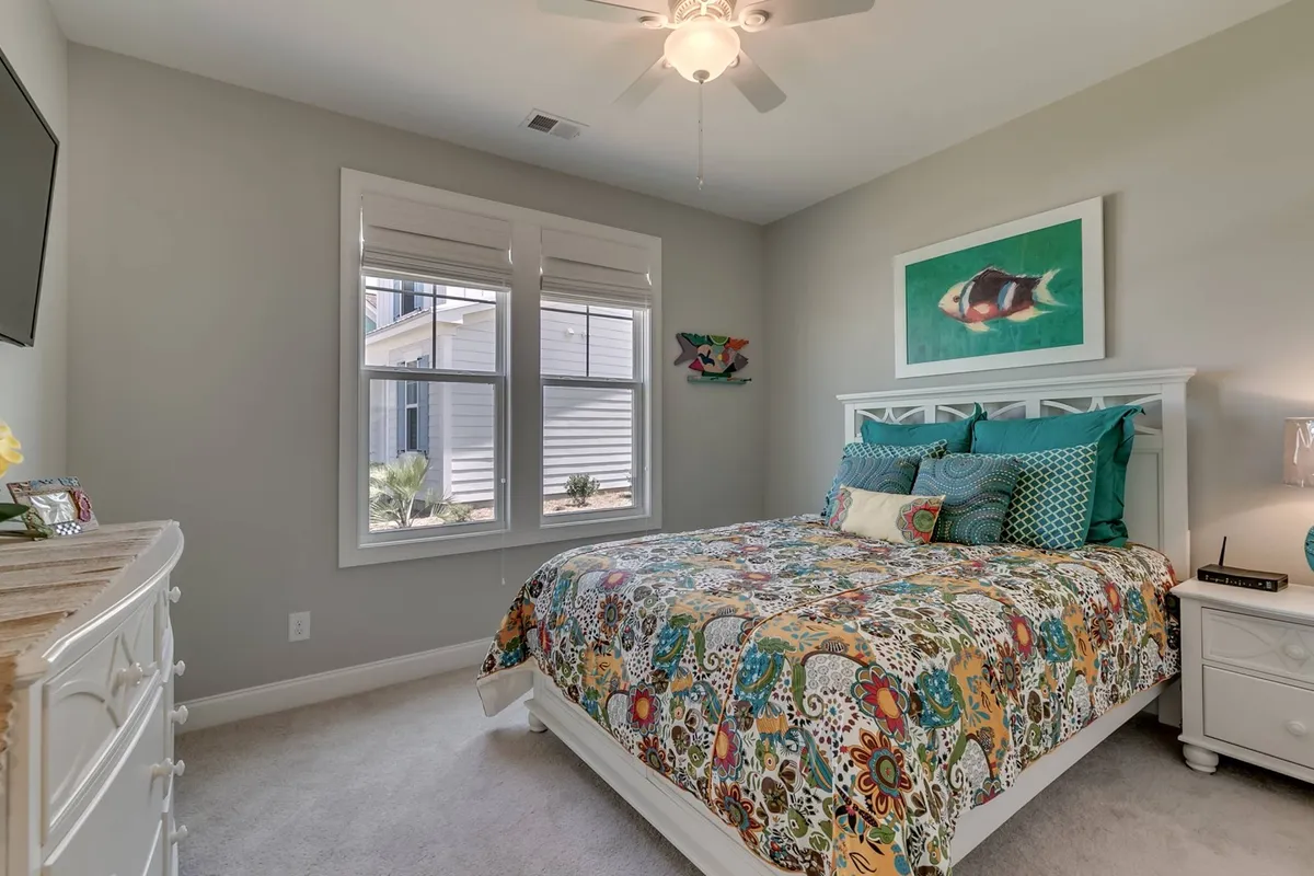 A cheerful bedroom with a colorful bedspread, framed fish artwork, and two windows that flood the space with natural light. The inviting decor and gentle ambiance highlight the room's fit for relaxation and rejuvenation.
