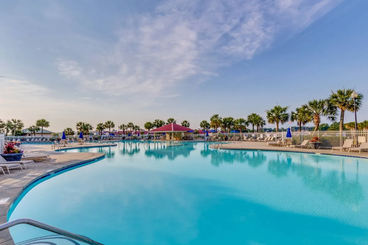 A vibrant exterior shot of the resort’s expansive swimming pool, surrounded by swaying palm trees, inviting poolside lounge chairs, and a cabana-style structure. The setting radiates relaxation and the joy of spending time outdoors with family and friends, with plenty of space for children to play and parents to unwind.