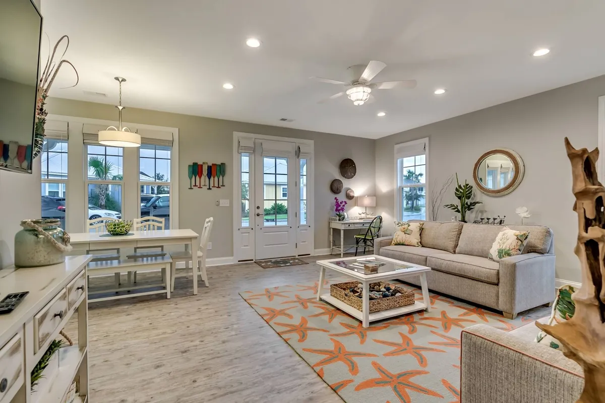 A wide view of the main living area showing a comfy sofa, a dining table large enough for family gatherings, decorative wall art, and large windows that flood the space with natural light. The overall feel is warm, inviting, and perfect for both relaxation and entertaining.