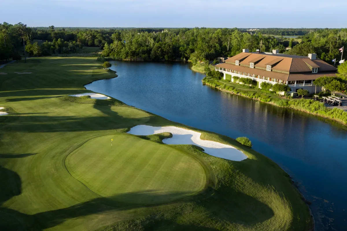 A sweeping view of a beautifully maintained golf course with a lake and a stately clubhouse in the background. Sand traps and lush greens set a scenic, inviting scene, perfect for golfers and those seeking a peaceful resort environment.