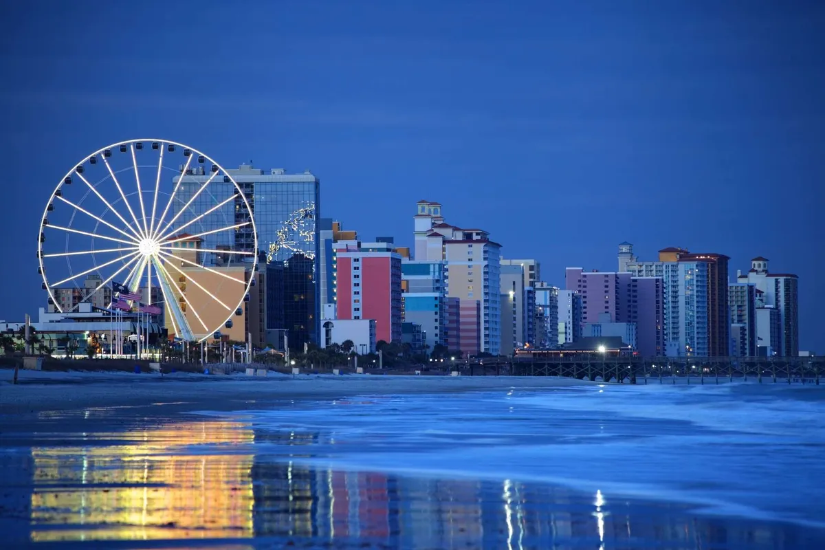 A vibrant scene featuring a Ferris wheel, colorful waterfront buildings, and the reflection of lights on the ocean. The area suggests a festive boardwalk atmosphere with opportunities for dining and entertainment near the beach.